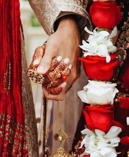 An Indian bride and groom's hands held together in traditional wedding attire with red and white roses
